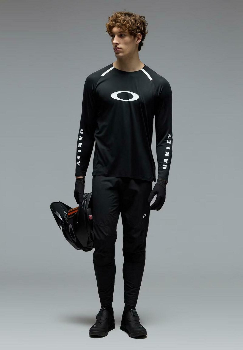 Jeune homme portant un shirt à manches longues Oakley noir, un pantalon et des gants, tenant un casque de moto noir, se tenant devant un fond gris.