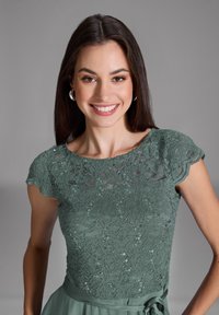 Grünes Spitzenkleid mit einem wellenförmigen Ausschnitt und kurzen Ärmeln, verziert mit Pailletten. Glatter Stoff, der an der Taille gerafft ist.