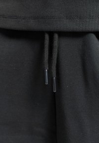 Ceinture en tissu noir avec deux cordons tressés pendants, agrémentés d'embouts en plastique aux extrémités.