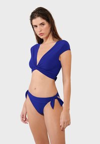 Dvodijelni bikini u vibrantnoj plavoj tkanini. Gornji dio ima kratke rukave i ispleteni prednji detalj. Donji dio ima nagomilane naglaske sa strane.