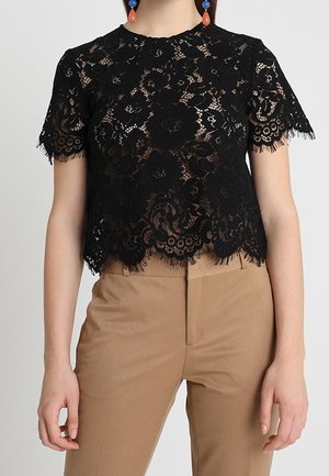 Woman wearing a black short-sleeve lace top with floral patterns and tan high-waisted pants against a plain background.