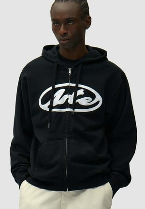 Giovane uomo che indossa una felpa nera con zip e un logo ovale bianco con testo sul petto, posando con le mani nelle tasche.