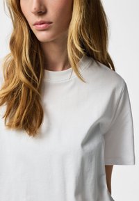 Femeie cu păr blond lung purtând un tricou alb simplu cu guler rotund, prezentată de la umeri până la bărbie pe un fundal alb.