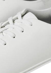 Des baskets en cuir blanc présentent une surface lisse, un bout rond et des lacets plats. Elles possèdent un design minimaliste avec une doublure grise.