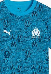 T-shirt turquoise à manches courtes avec un motif répété de logos marseillais bleu foncé et le texte "Droit au But". Texture de tissu douce.