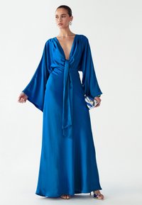 Lange blauwe satijnen jurk met wijde mouwen, diepe V-hals en een strik in de taille. Model draagt een zilveren clutch en oorbellen met steentjes. Tot op de grond reikend.