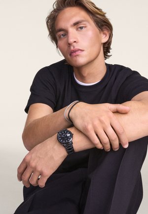 Orologio da polso nero con quadrante rotondo, rifiniture argentate e cinturino a maglie nere; indossato su un polso con un bracciale grigio. Modello con una t-shirt nera.