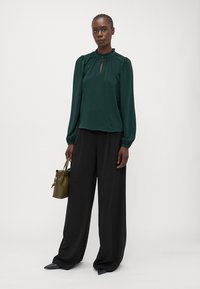Groene blouse met lange mouwen en hoge halslijn, met een keyhole-detail aan de voorkant en geplooide schouders, gecombineerd met zwarte ruime broek en een kleine olijfgroene tas.