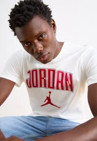 Biela bavlnená tričko s nápisom "JORDAN" v tučných červených písmenách a ikonickým logom Jordan v červenej farbe, s krátkymi rukávmi a klasickým strihom.