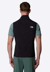 Black vest with a high collar, made of lightweight fabric, features a logo on the back and is paired with a short-sleeve, teal shirt.