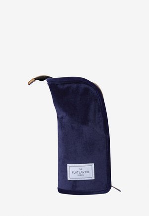 Mörkblå sammet kosmetikapåse med en texturerad yta, utrustad med ett guldzipper och en vit etikett med texten "THE FLAT LAY CO. LONDON."