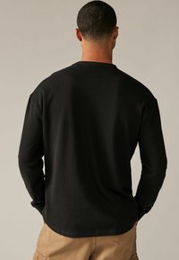 Chemise noire à manches longues avec un tissu texturé, un col rond et une coupe décontractée. Le dos présente une finition lisse et des coutures discrètes.