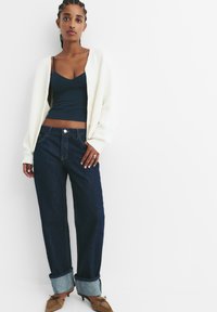 Blaue Jeans mit aufgerollten Säumen, kombiniert mit einem dunkelblauen Tanktop und einem übergroßen weißen Rippstrick-Cardigan. Braune Absätze runden das Outfit ab.
