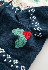 Granatowy sweter z dzianiny z białymi wzorami i szydełkową dekoracją w postaci ostrokrzewu w kolorach zielonym i czerwonym na kieszeni.