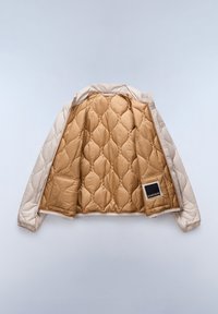 Donsjack met een beige buitenkant en een gouden gewatteerde binnenkant, voorzien van een patch pocket en een ritssluiting. Zachte textuur en gladde voering.