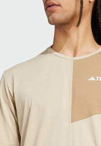 Beige short-sleeve t-shirt featuring a contrasting tan panel and white logo. Smooth fabric with a round neckline and regular fit.