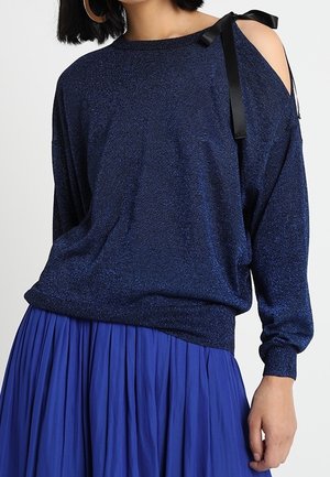 Frau trägt einen dunkelblauen schulterfreien Pullover mit schwarzem Banddetail und einen plissierten königsblauen Rock, stehend vor einem einfarbigen Hintergrund.