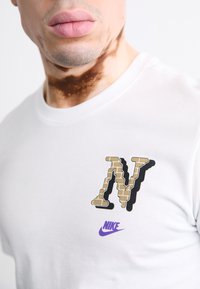 Witte katoenen T-shirt met een gestructureerd zwart en gouden "N" logo en een paarse Nike swoosh, met een ronde hals ontwerp.