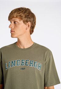 Olivegrön bomullströja med rundad hals och korta ärmar, med "LINDBERGH 1927" broderat i marinblått och vitt.