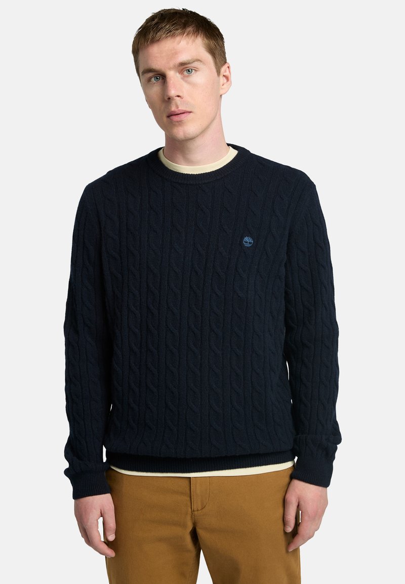 Maglione lavorato a maglia navy con texture a trecce, polsini e orlo a coste, caratterizzato da un piccolo logo blu sul petto. Indossato sopra una camicia chiara.