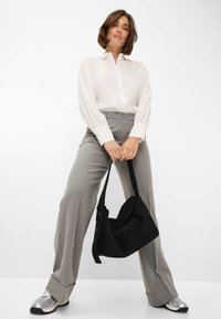 Zwarte suède handtas met een gebogen vorm, crossbody gedragen. Model in een wit shirt en grijze high-waisted broek, met zilveren sportschoenen.