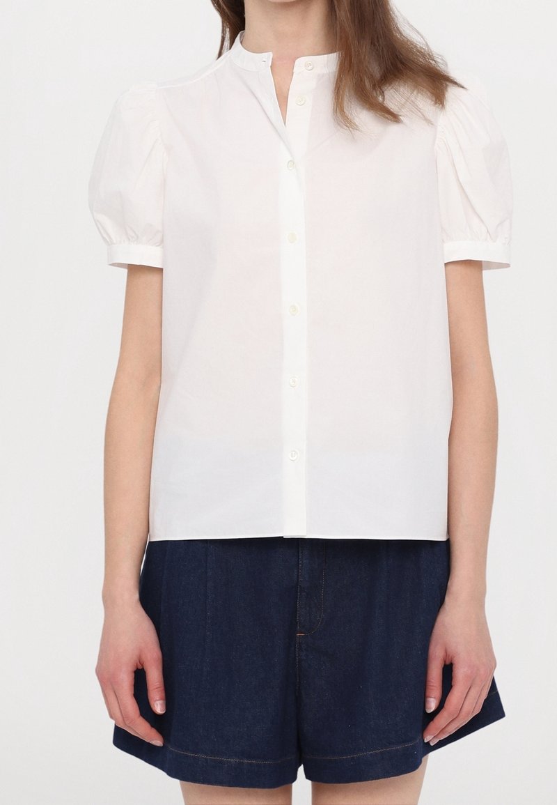 Blouse blanche à manches courtes boutonnée avec manches bouffantes portée avec un short taille haute bleu marine foncé.