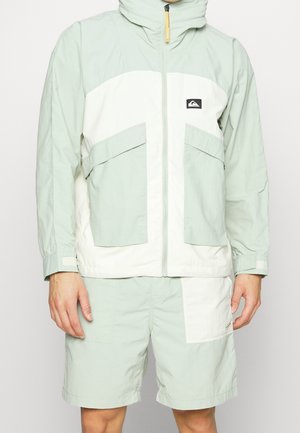 Man wearing light green and white zip-up jacket with large front pockets and matching shorts with a white patch on one leg.