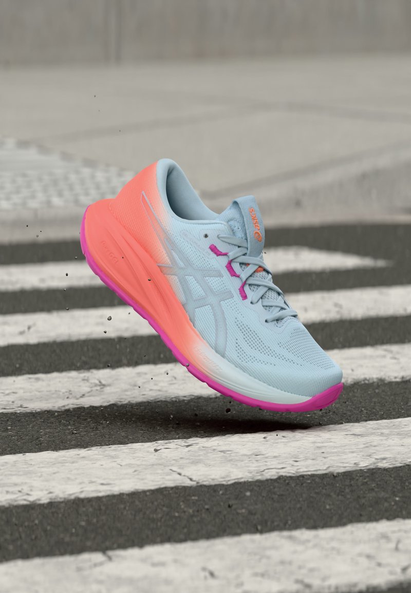 Zapatilla de correr azul claro y rosa en el aire sobre un paso de peatones en una calle asfaltada, con pequeños escombros levantados alrededor de la suela.