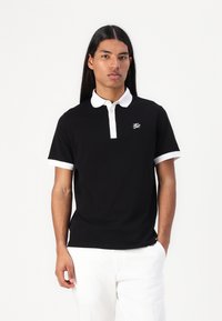 Μαύρο polo μπλουζάκι με λευκό γιακά και μανίκια, διαθέτει κουμπωτή σειρά και μικρό λογότυπο στο στήθος. Λειαντική υφή υφάσματος.