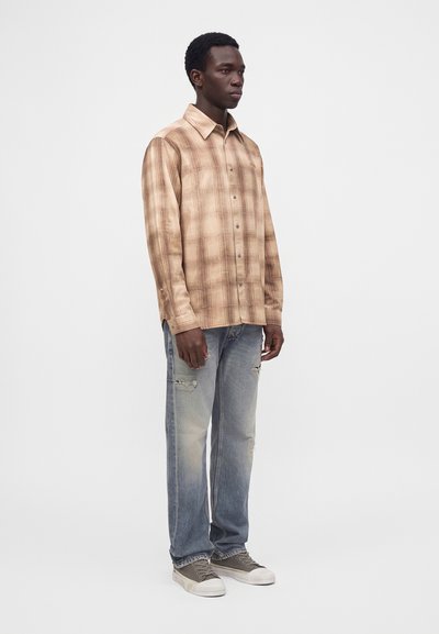 Homme portant une chemise à carreaux beige à manches longues, un jean bleu délavé et déchiré, et des baskets grises, debout sur un fond blanc uni.