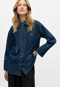 Camicia in jeans scuro con colletto a papillon, tasca frontale e logo dorato. Presenta maniche lunghe e una vestibilità rilassata con orlo leggermente curvato.