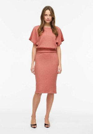 Kvinne som står vendt fremover iført en støvet rosa strikket kortermet topp og matchende knelang blyantskjørt med svarte åpne tåhøye hæler.