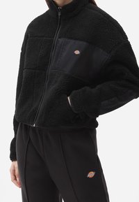 Giacca nera in fleece con pannelli trapuntati e chiusura con zip. Presenta una patch con il logo sul petto e un collo alto. Texture morbida e vestibilità rilassata.