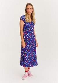 Žena v modrých midi šatech se barevnými vzory ovoce a zeleniny, v růžových teniskách, stojící na čistě bílém pozadí.