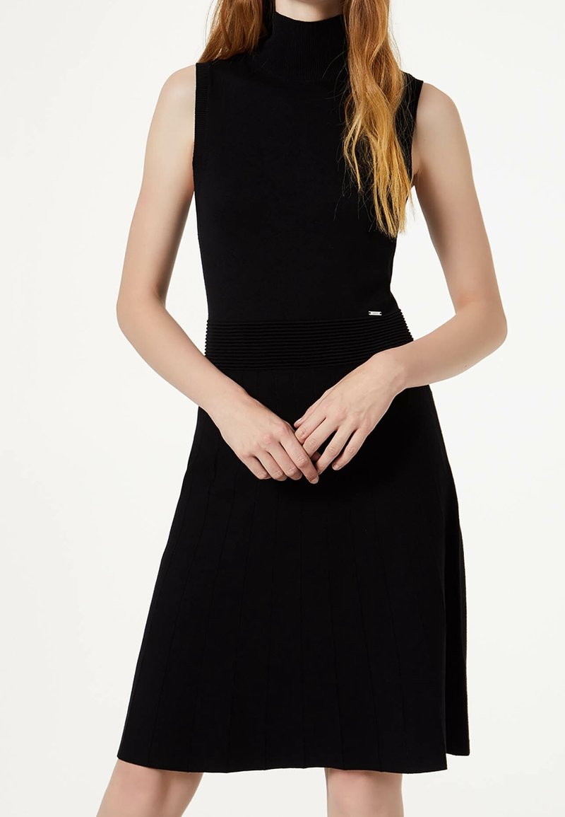 Femme portant une robe noire sans manches à col montant et taille côtelée, debout les mains jointes au niveau du ventre sur un fond blanc.