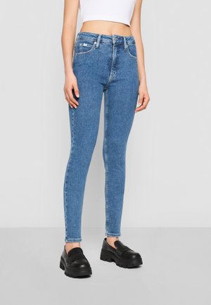 Femme portant un jean skinny bleu taille haute, un top court blanc et des chaussures plateformes noires épaisses sur un fond uni.
