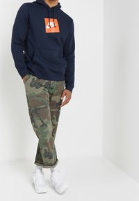 Marinblå huvtröja med orange Nike-logga, kombinerad med gröna kamoflagebyxor och vita sneakers. Avslappnad look med kontrasterande färger och texturer.