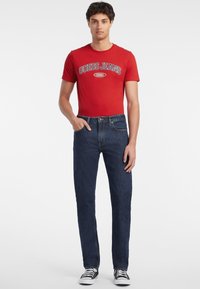 Tricou roșu cu logo-ul "GUESS JEANS 1981", asortat cu blugi de denim de culoare albastru închis. Modelul poartă adidași negri și albi, stând cu o mână în buzunar.