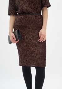 Brūns sekvencēts kleita ar pieguļošu siluetu, īsām piedurknēm, jostotu vidukli, apvienota ar melnu clutch somu un necaurspīdīgām melnām zeķbiksēm.