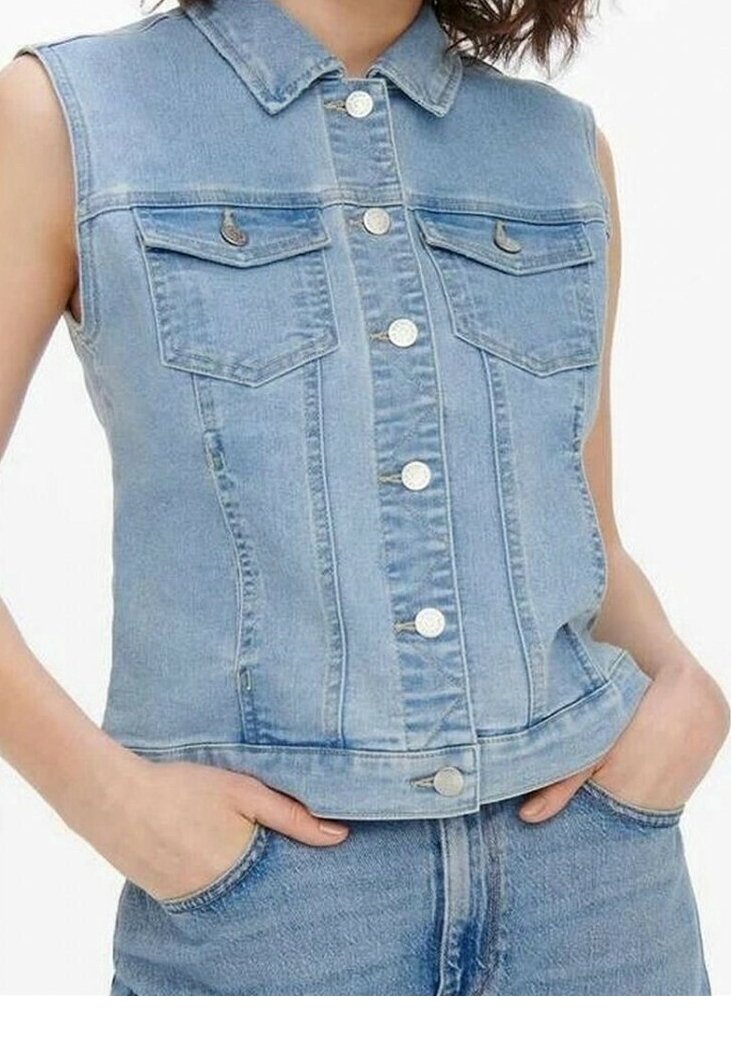 Veste en jean sans manches bleu clair avec des boutons argentés, deux poches poitrine, portée sur un jean assorti avec les mains dans les poches.