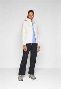 Giacca leggera in crema con cappuccio, abbinata a una felpa blu e pantaloni cargo neri. Le scarpe sono grigie con suole testurizzate.