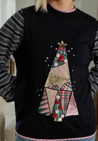 Schwarzer Pullover mit einem Weihnachtsbaum-Design, versehen mit bunten Stoffen, Pailletten und Bommel. Gemusterte Ärmel und ein geschwungener Halsausschnitt.