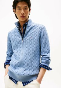 Hellblauer, kabelgestrickter Zip-Pullover mit hohem Kragen, über einem navy- und weißen gestreiften Hemd getragen, kombiniert mit cremefarbenen Hosen.