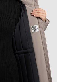 Padded bunda s béžovým exteriérom a čiernym interiérom, s priehlbínovým textúrovým vzorom, prešívaním a etiketou s nápisom: "Keď nájdeš dobré oblečenie..."