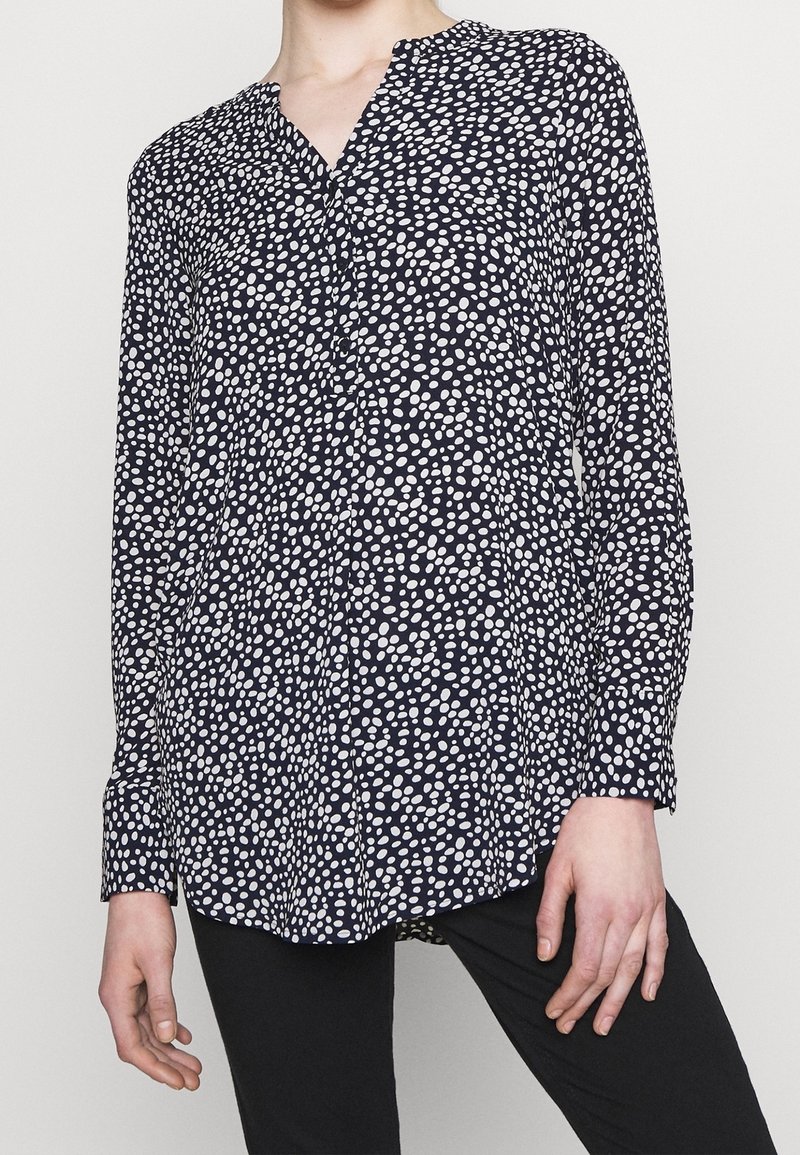 Femme portant un chemisier bleu marine à manches longues avec des pois blancs irréguliers, associé à un pantalon noir, debout devant un fond clair uni.