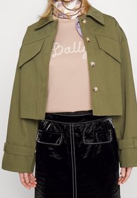 Personne portant une veste vert olive avec des boutons, une chemise rose clair avec un texte manuscrit, une jupe noire brillante avec des poches et un foulard à motifs autour du cou.