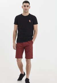 Camiseta de algodón negra con un pequeño gráfico en rojo y azul, combinada con pantalones cortos rojos. El modelo lleva zapatillas negras y adopta una pose neutral.