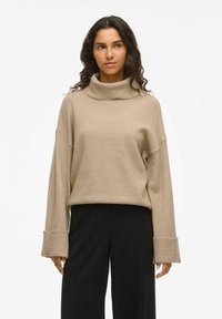 Bežna, mehka pletena turtleneck pulover z spuštenimi rameni, širokimi rebrastimi manšetami in sproščenim krojem, v kombinaciji s črnimi širokimi hlačami.