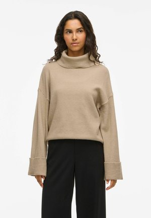 Pull à col roulé beige en tricot doux avec épaules tombantes, larges poignets côtelés et coupe décontractée, associé à un pantalon noir à jambes larges.