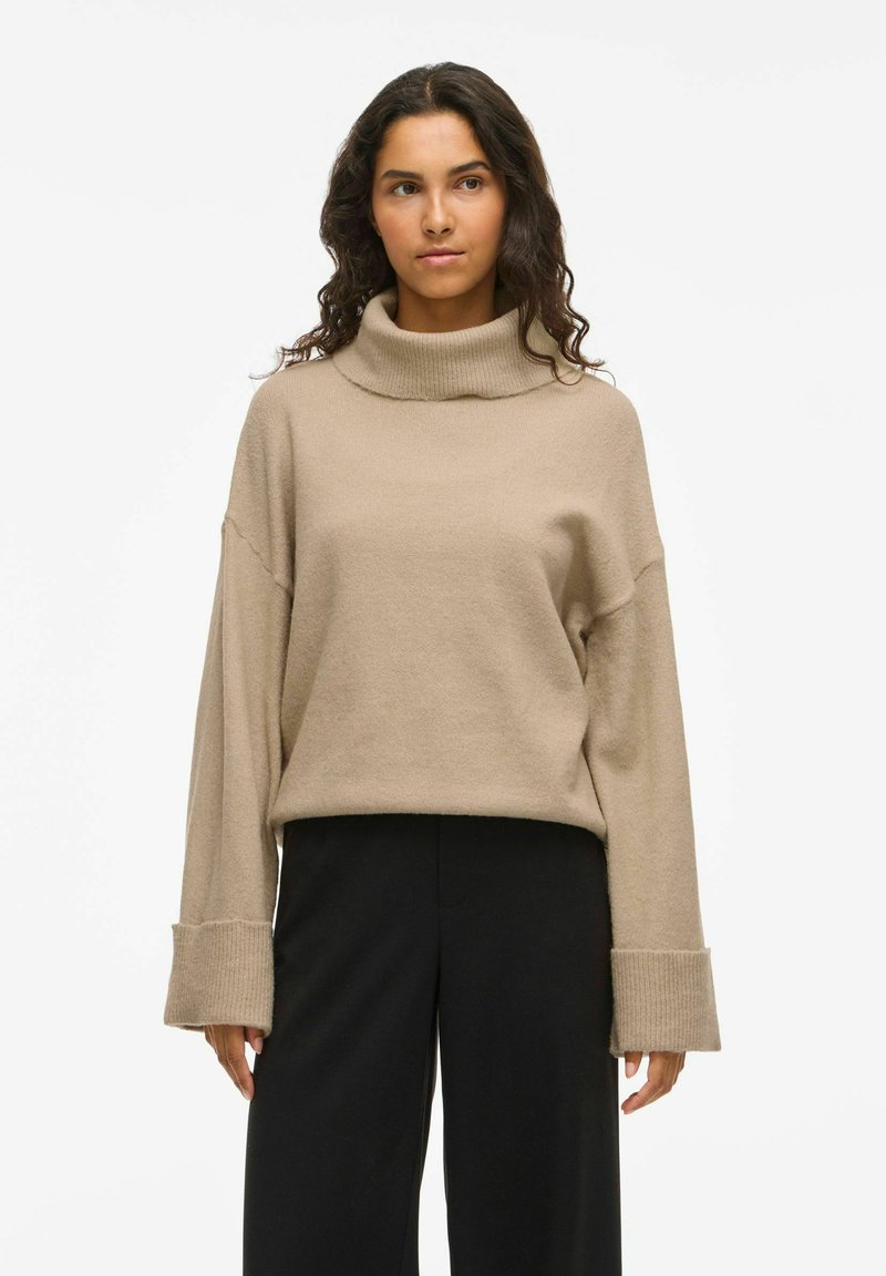 Bežna, mehka pletena turtleneck pulover z spuštenimi rameni, širokimi rebrastimi manšetami in sproščenim krojem, v kombinaciji s črnimi širokimi hlačami.